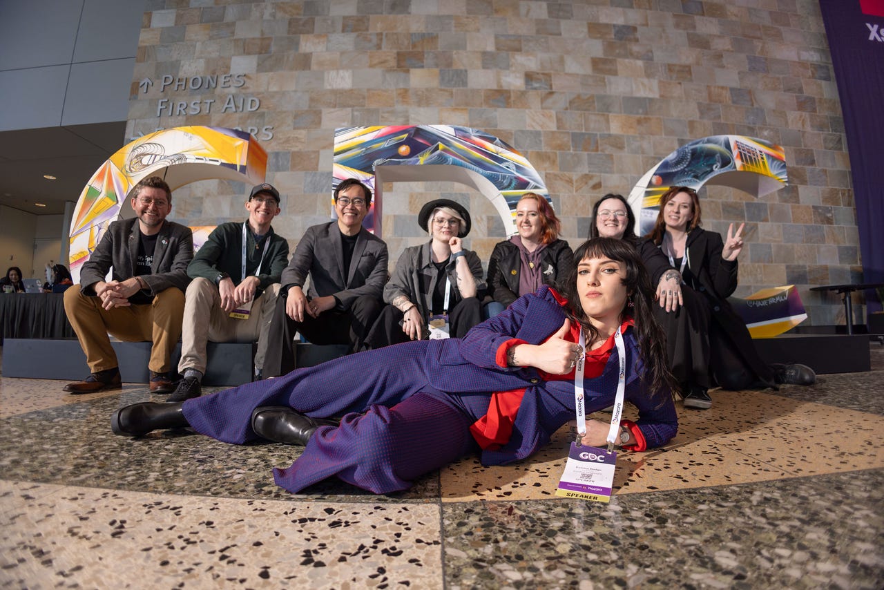 A group of people pose in front of the GDC logo, one woman in a blue suit lounges in front of the rest.
